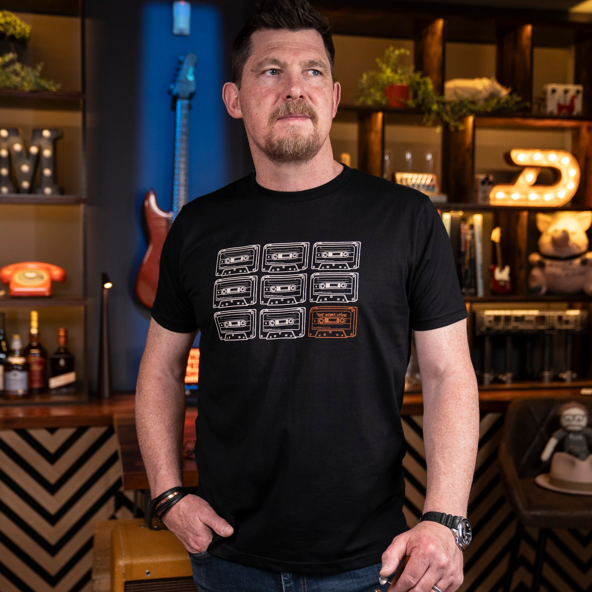 Mick from That Pedal Show wearing a black crewneck t-shirt with a graphic of nine cassette tapes in a 3x3 grid, called 'Mick's Tape'. Photographed in the bar studio at TPS HQ.