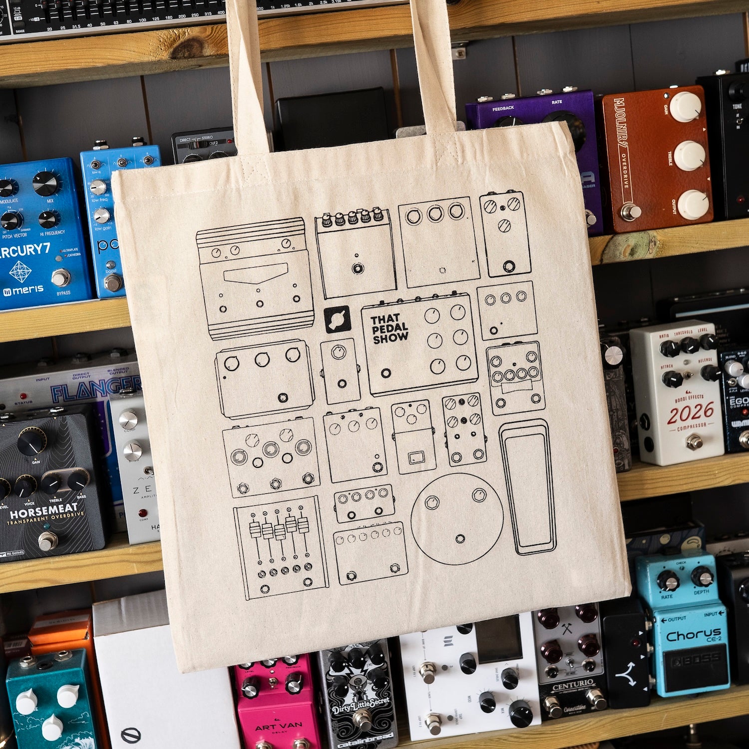 A natural canvas tote bag with black line drawings of guitar effects pedals, featuring the text 'THAT PEDAL SHOW' hanging in front of a wooden shelf filled with guitar effects pedals.