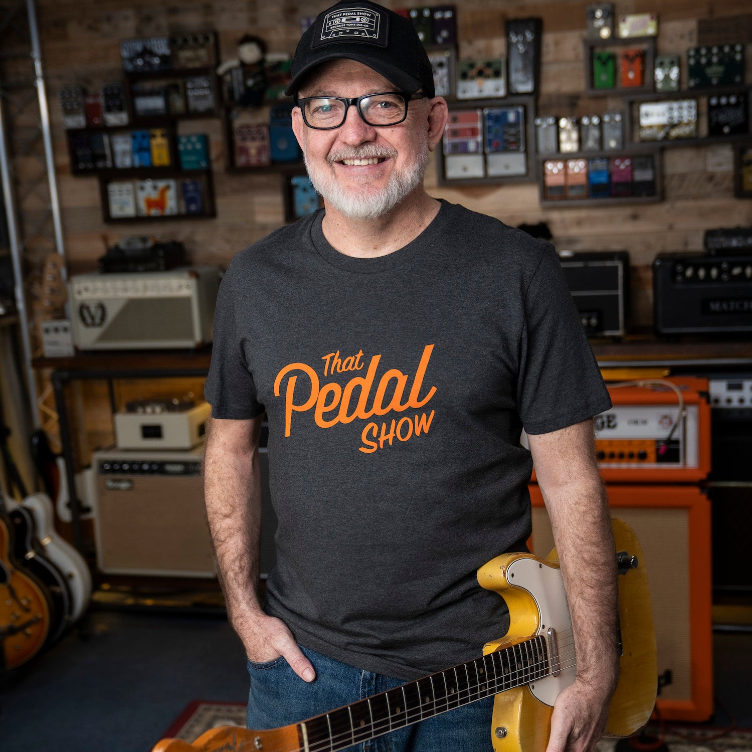 Dan wearing a 'That Pedal Show' vintage logo t-shirt holding a guitar in the TPS Studio