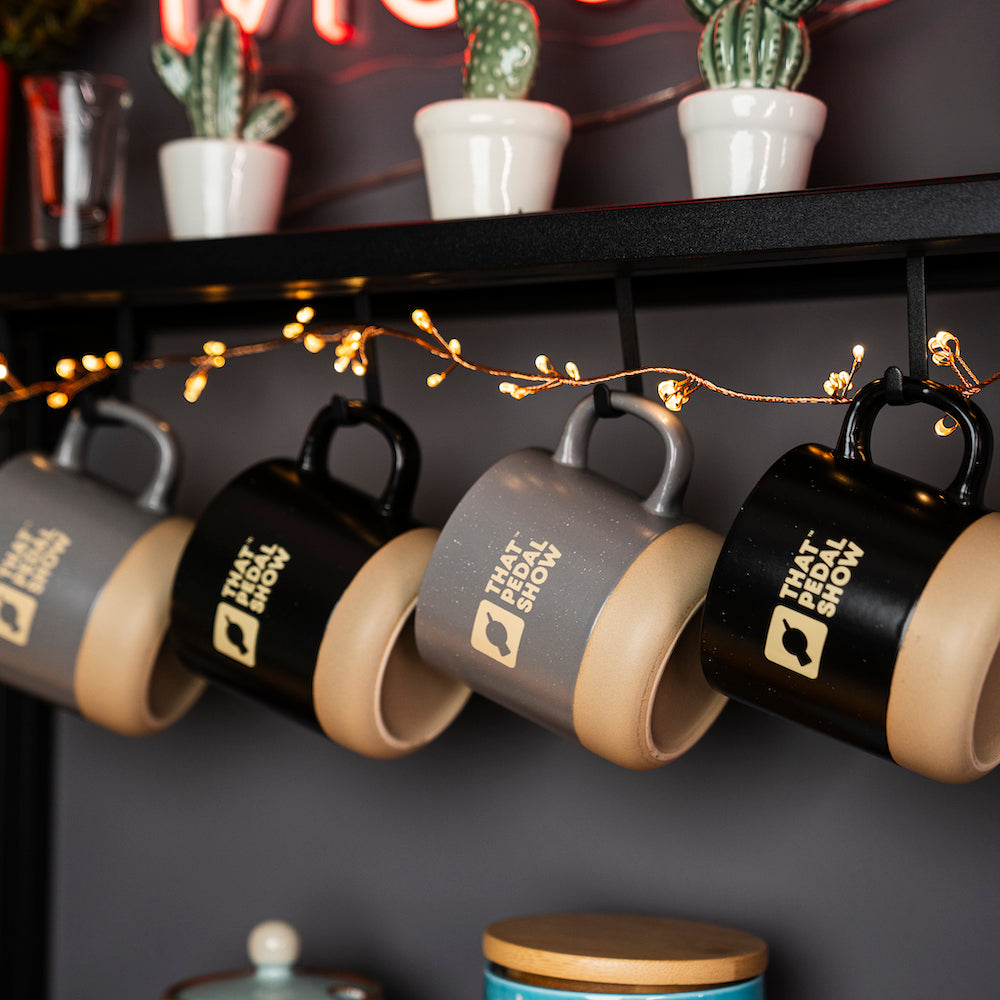 A set of That Pedal Show ceramic mugs with a speckled black and grey finish, hanging on a wall-mounted rack, with decorative lighting and plants in the background.
