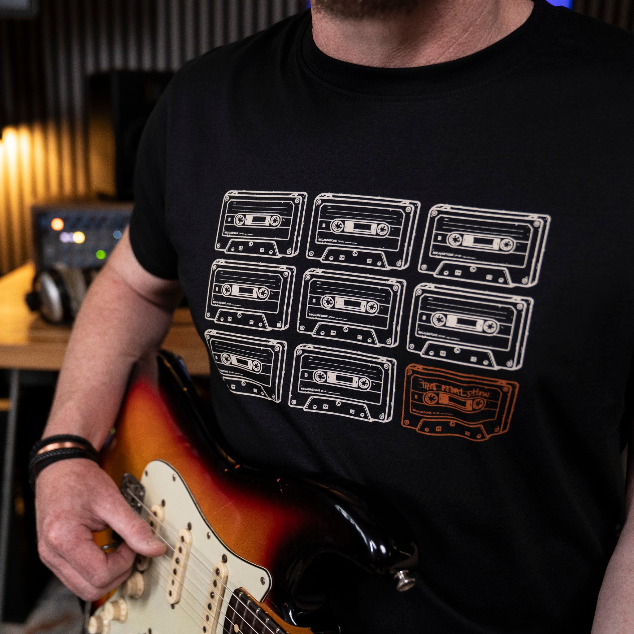 Mick Taylor wearing a black graphic t-shirt with a white and orange cassette tape print, holding a sunburst electric guitar in a studio setting.