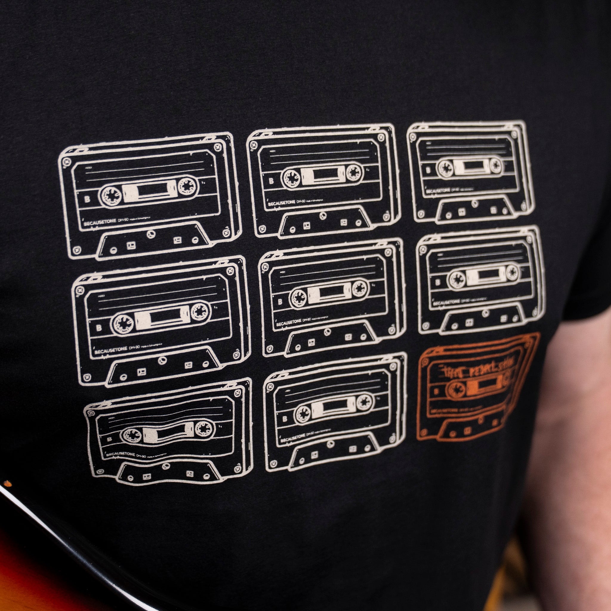 Close-up of a black T-shirt with a graphic print of nine cassette tapes, including one highlighted cassette in a dark orange colour, worn by Mick from That Pedal Show who is holding his Fender Stratocaster guitar.