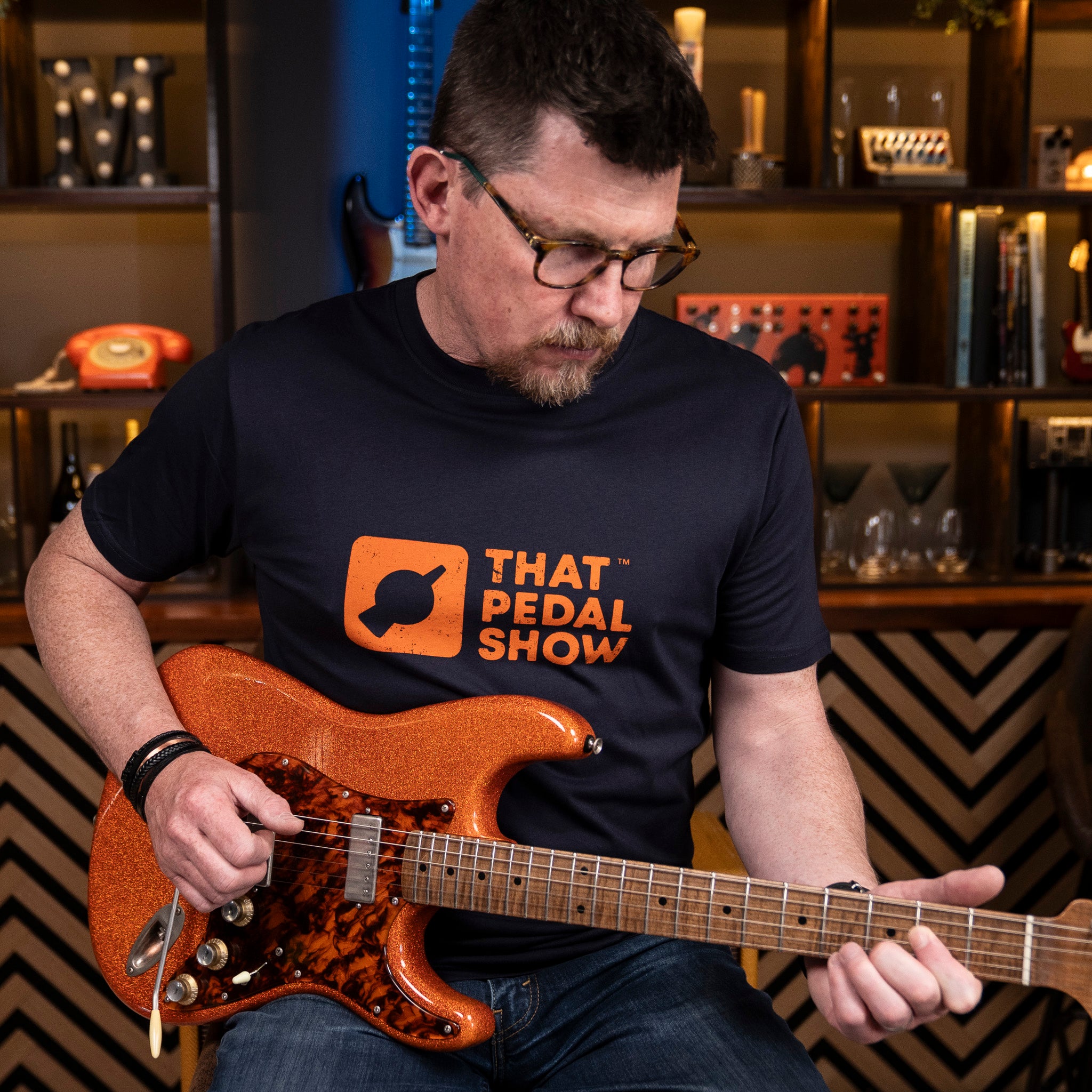 Mick from TPS wears a Navy blue t-shirt with the That Pedal Show logo printed in orange on the chest while holding his orange sparkle Cutler guitar