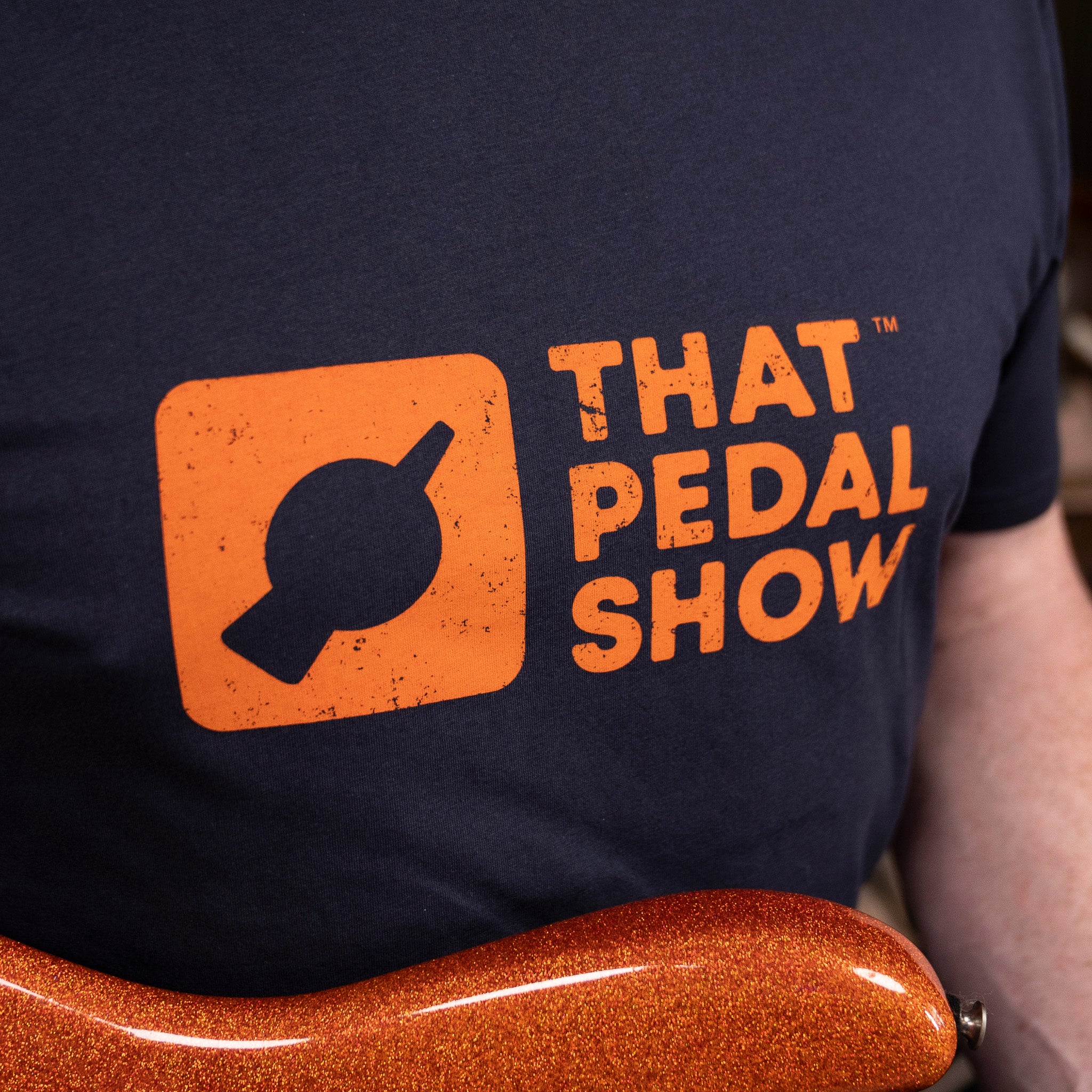 Mick from TPS wears a Navy blue t-shirt with the That Pedal Show logo printed in orange on the chest while holding his orange sparkle Cutler guitar