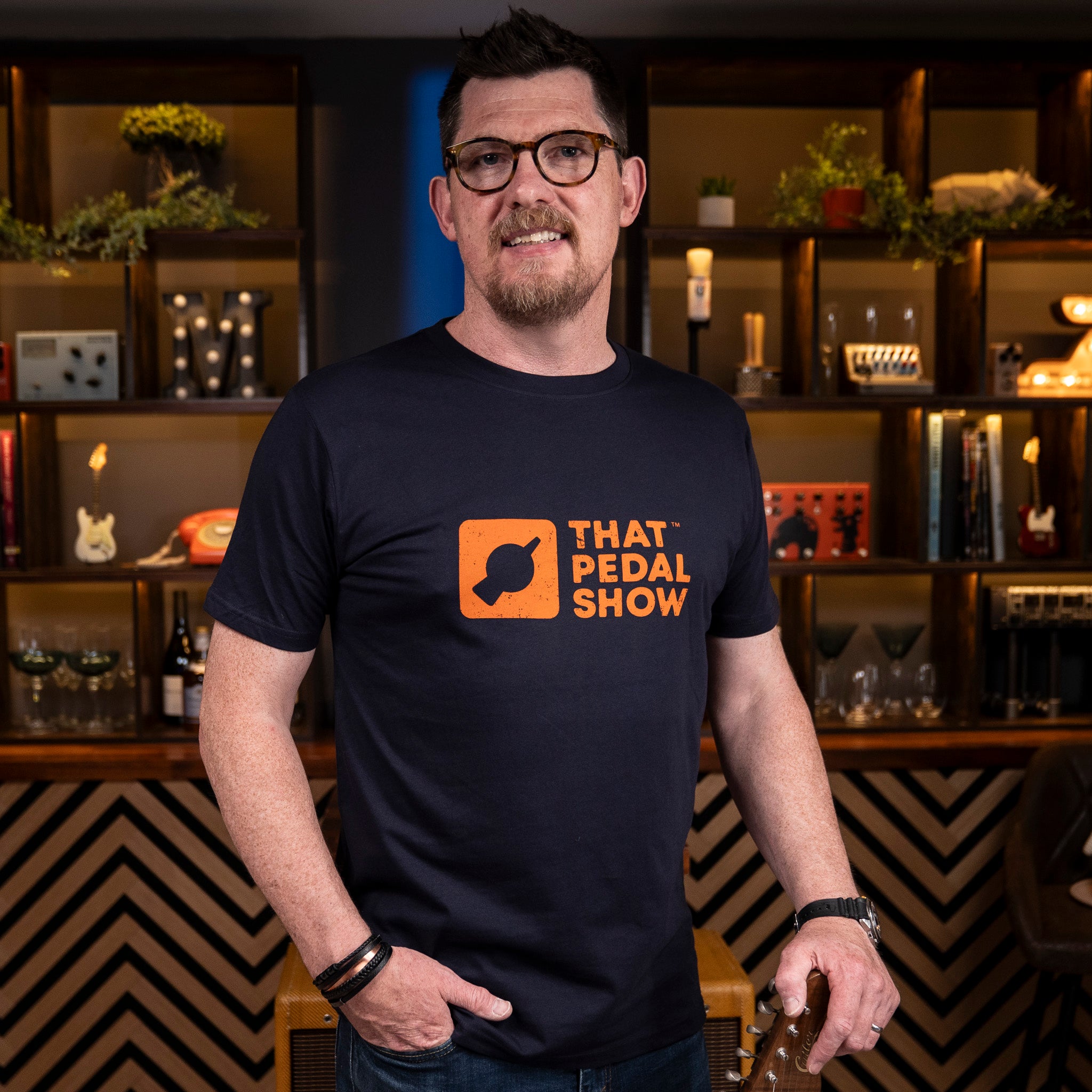 Mick from TPS wears a Navy blue t-shirt with the That Pedal Show logo printed in orange on the chest. Photographed in the 'Bar' studio at TPS HQ