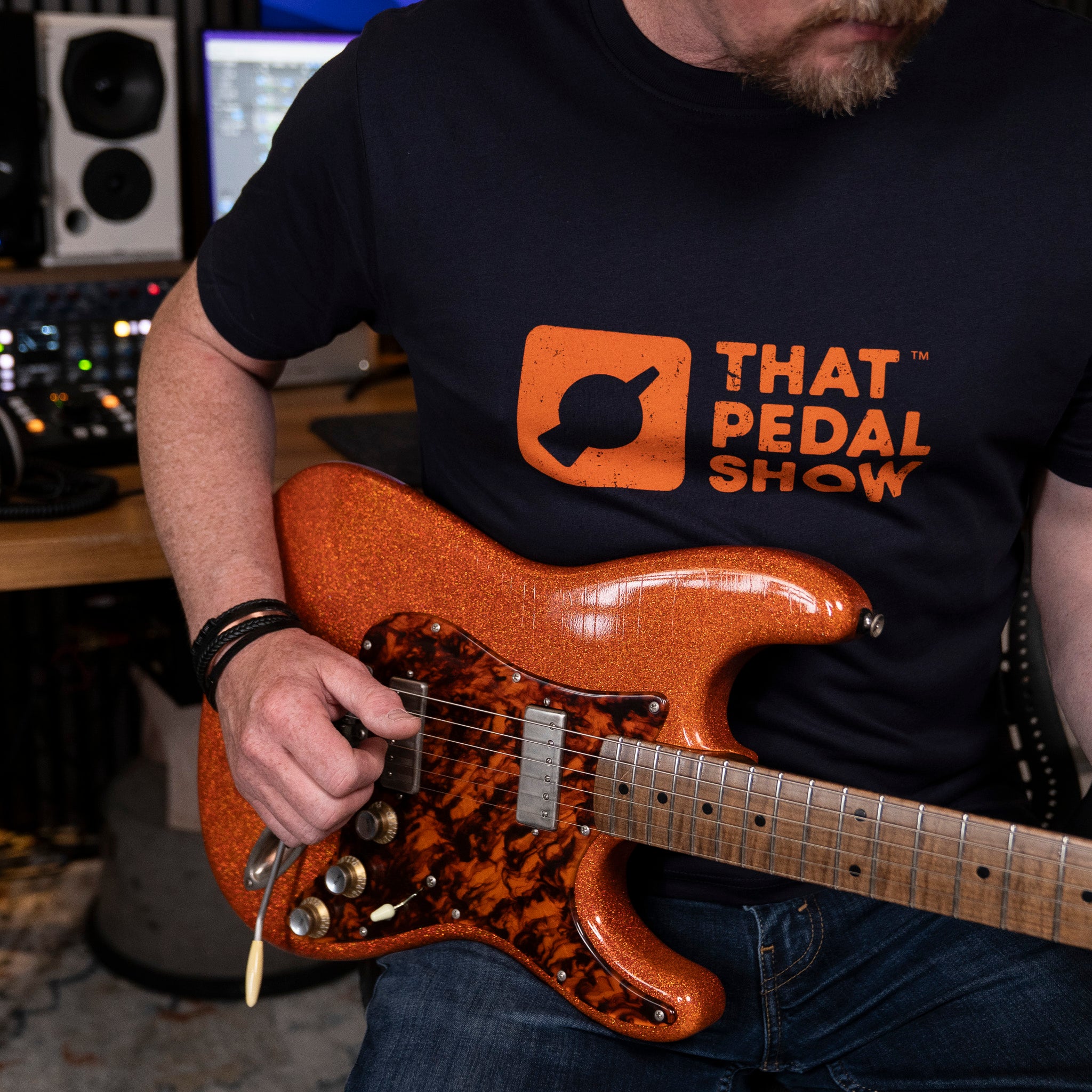 Mick from TPS wears a Navy blue t-shirt with the That Pedal Show logo printed in orange on the chest while holding his orange sparkle Cutler guitar