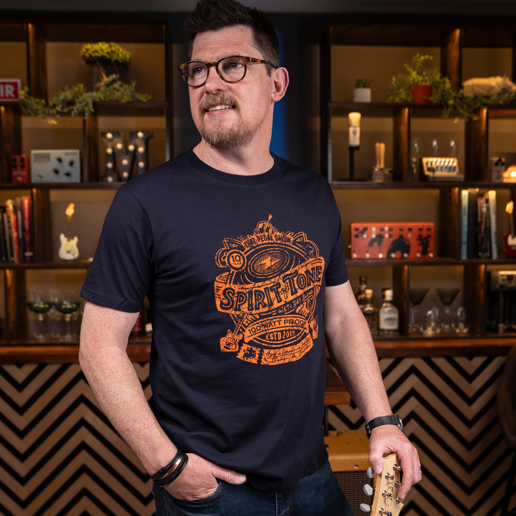 Mick from That Pedal Show wears the 'Spirit of Tone' t-shirt design in orange on a navy blue tee. Photographed in the Schwangri-Bar Studio at TPS HQ
