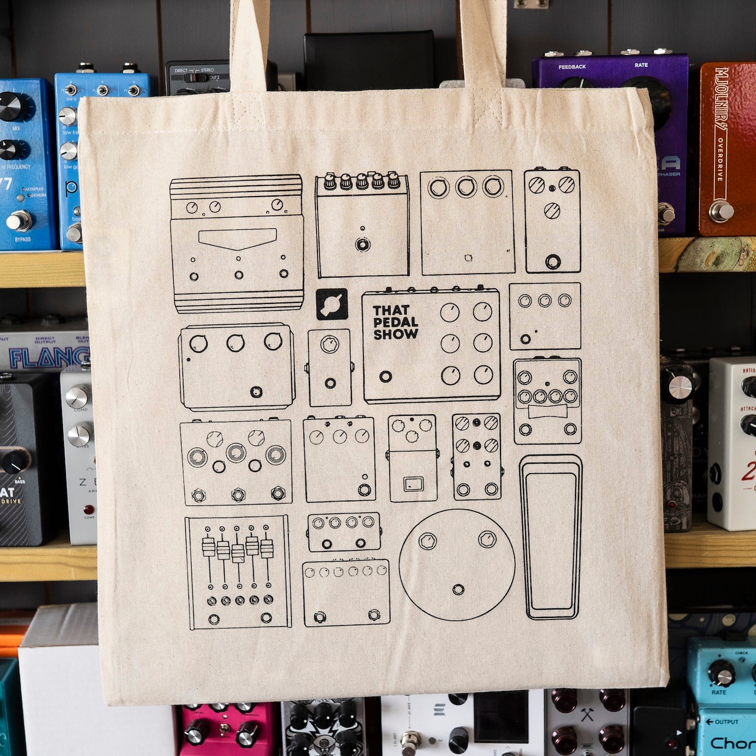 A natural canvas tote bag featuring black line-art illustrations of guitar effects pedals, with the text 'THAT PEDAL SHOW' printed on one of the pedal boxes. The bag is hanging in front of a wooden shelf filled with colourful guitar effects pedals.