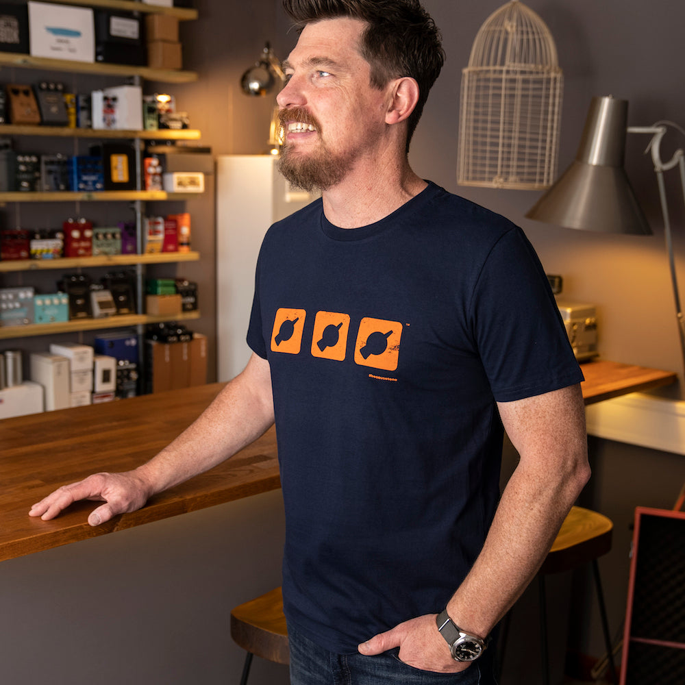 Mick from That Pedal Show wearing a navy t-shirt with the 'Three Dials' design featuring the TPS chicken head 'knobicon' logo in orange.
